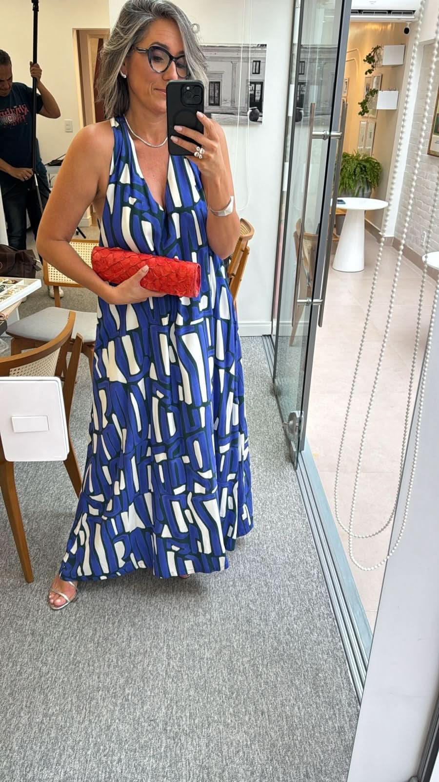 Mulher usando vestido longo azul estampado, segurando clutch vermelha em ambiente moderno EZILDINHA