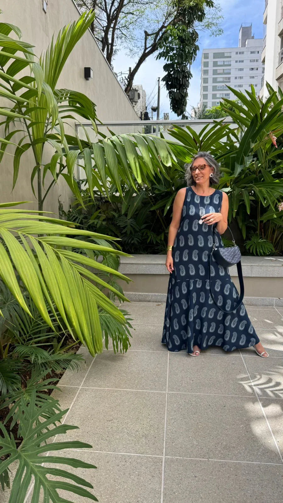 Mulher usando vestido longo azul de viscose estampado, ao ar livre, cercada por plantas tropicais EZILDINHA