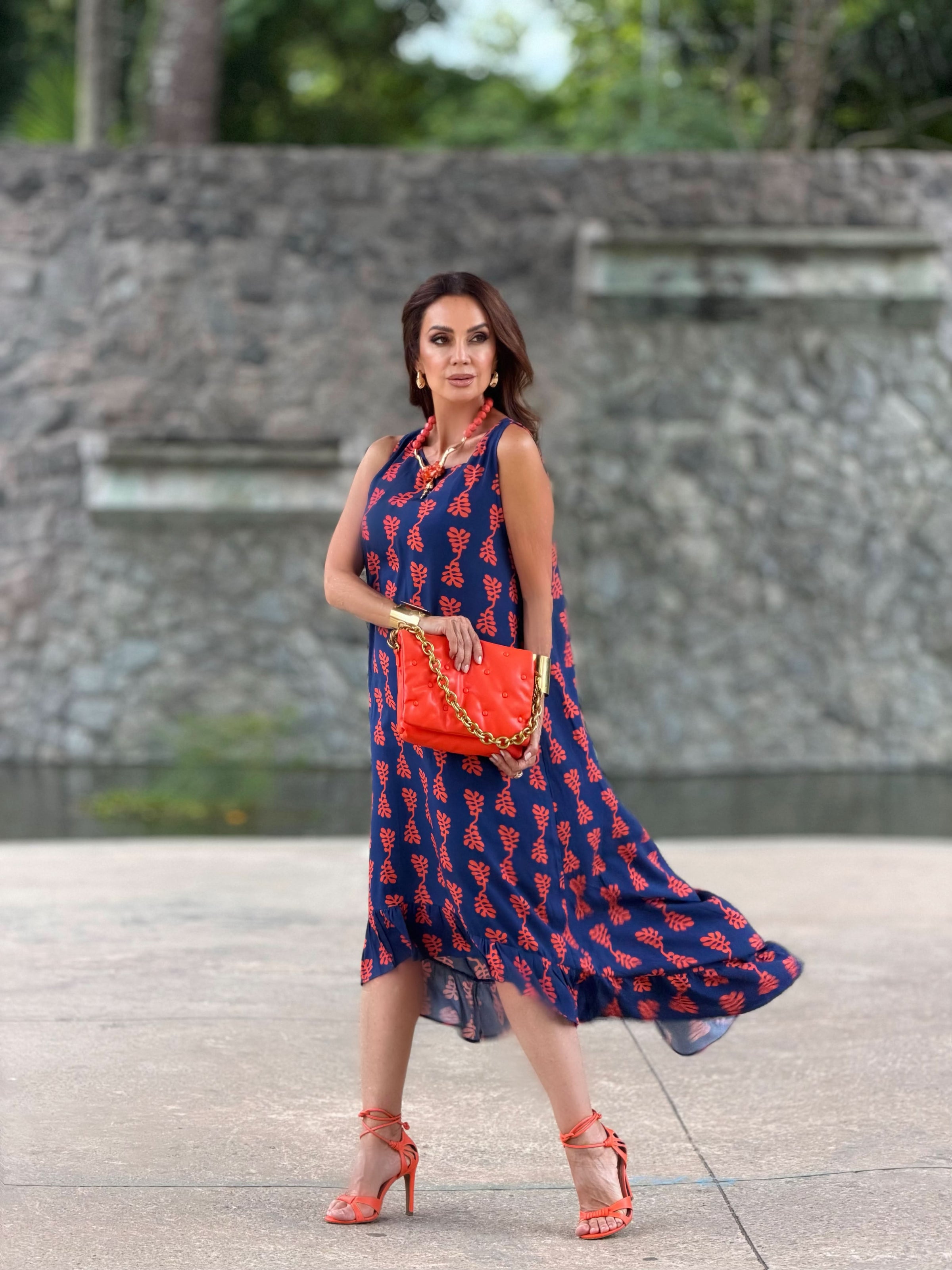 Mulher usando vestido midi azul estampado com folhas vermelhas, bolsa e sandália laranja, ao ar livre EZILDINHA