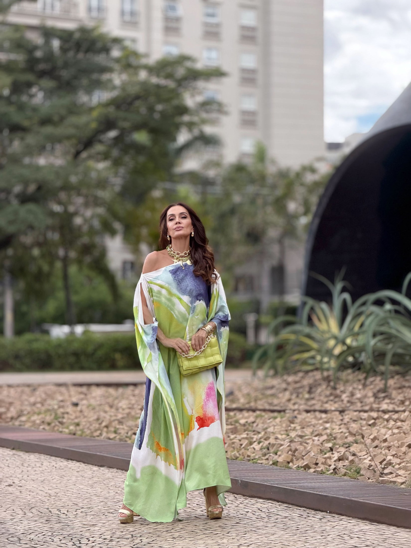Mulher usando vestido kaftan longo estampado colorido ao ar livre, bolsa verde, sandálias plataforma EZILDINHA