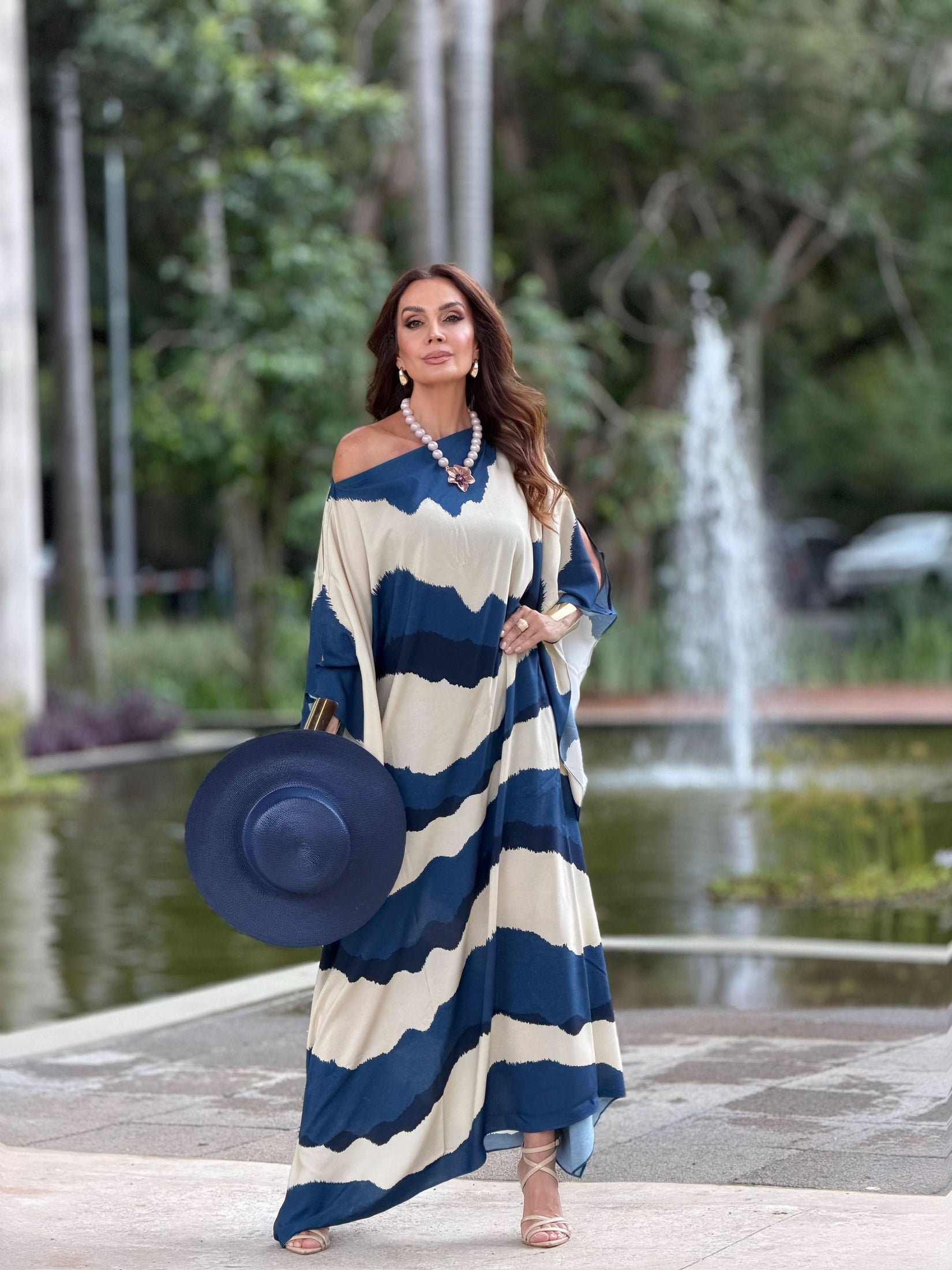 Mulher usando vestido kaftan longo azul e bege, segurando chapéu, ao ar livre junto a fonte EZILDINHA