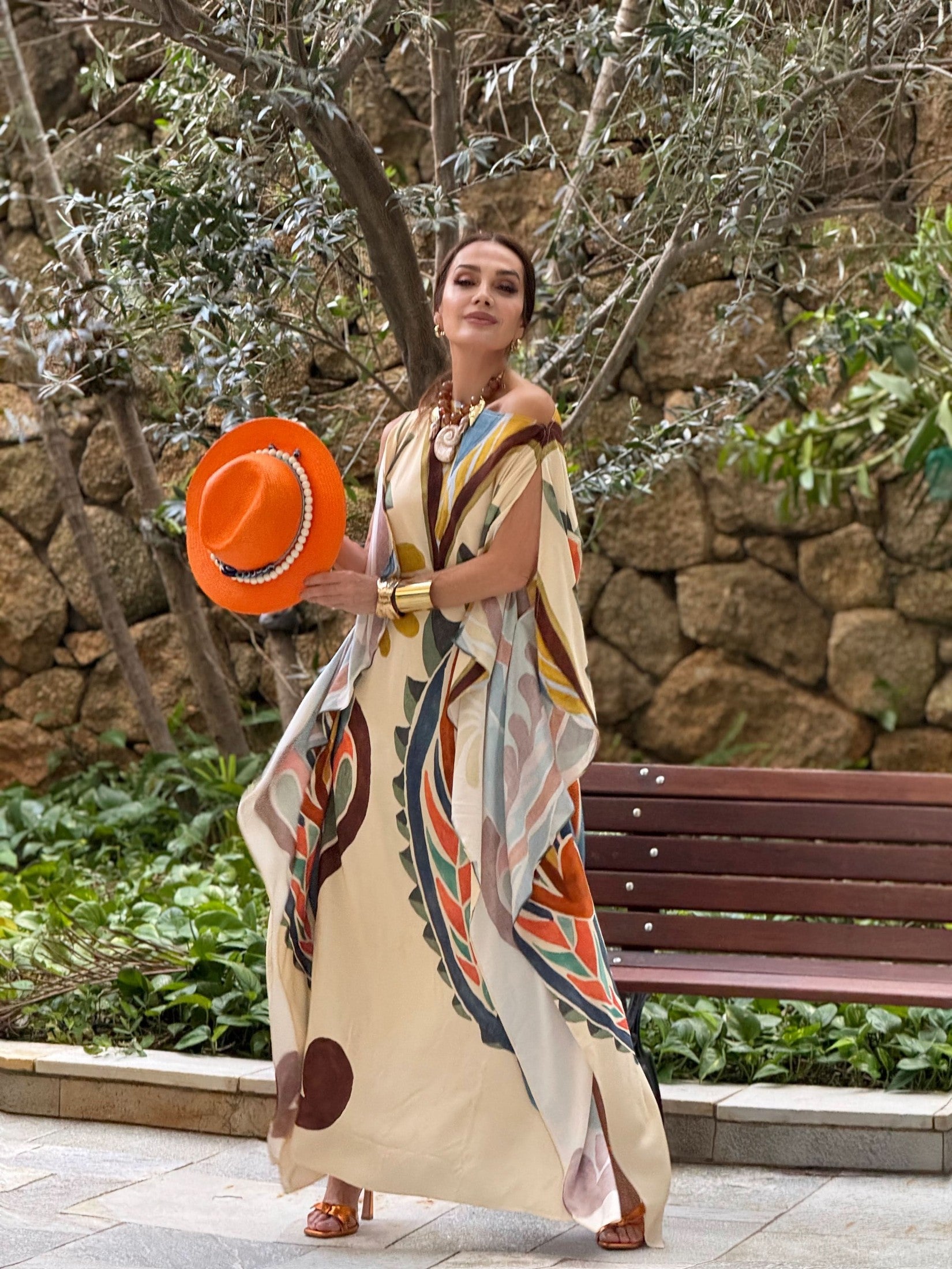 Mulher usando vestido kaftan estampado, segurando chapéu laranja em jardim com bancos EZILDINHA