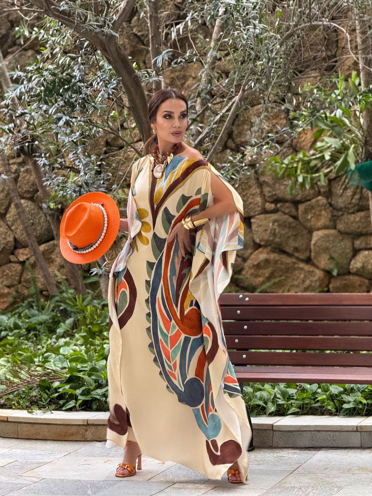 Mulher usando vestido kaftan estampado longo em seda, segurando chapéu laranja, em jardim EZILDINHA
