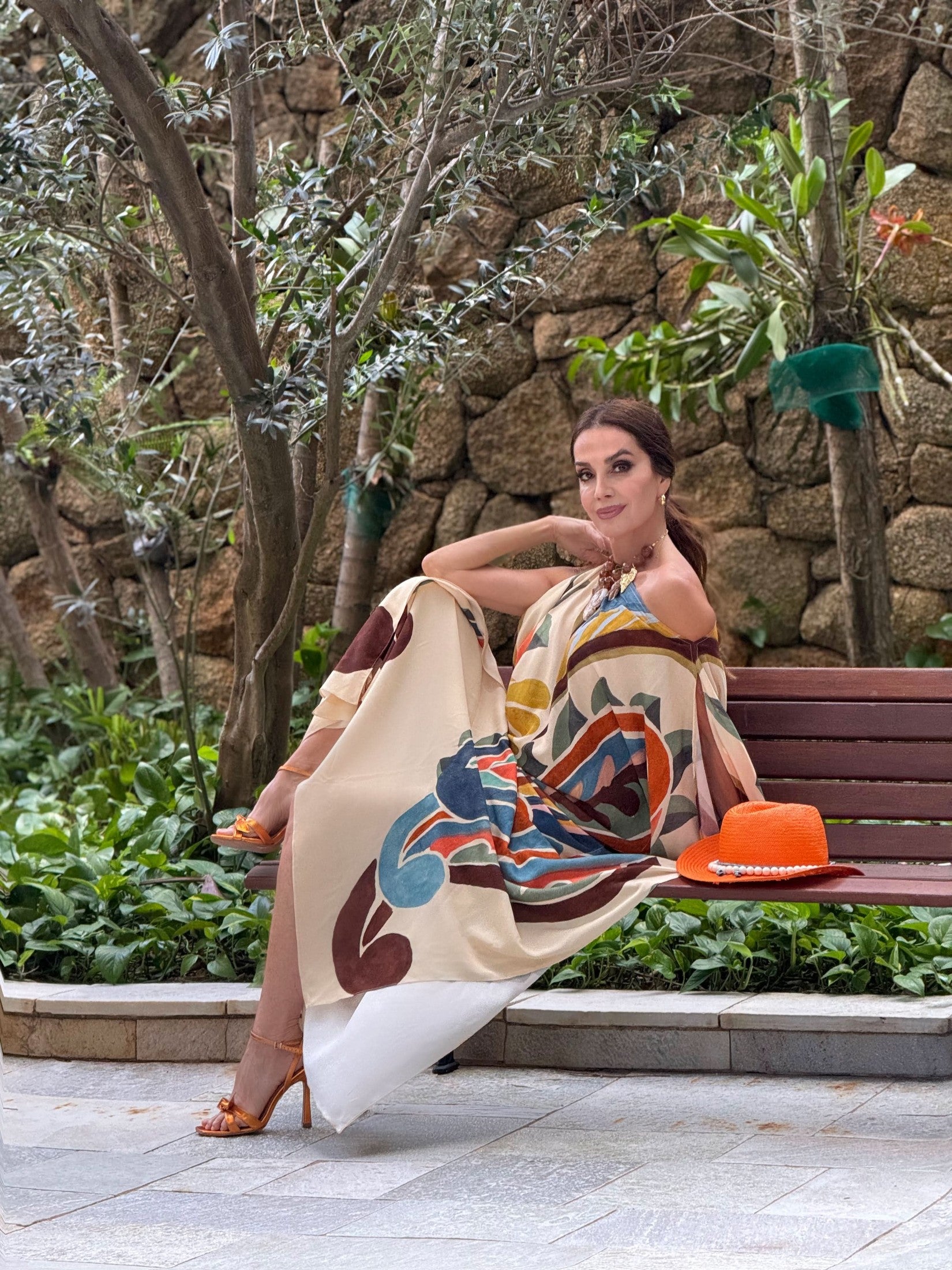 Mulher usando vestido kaftan estampado, sentada em banco de jardim com chapéu laranja ao lado EZILDINHA
