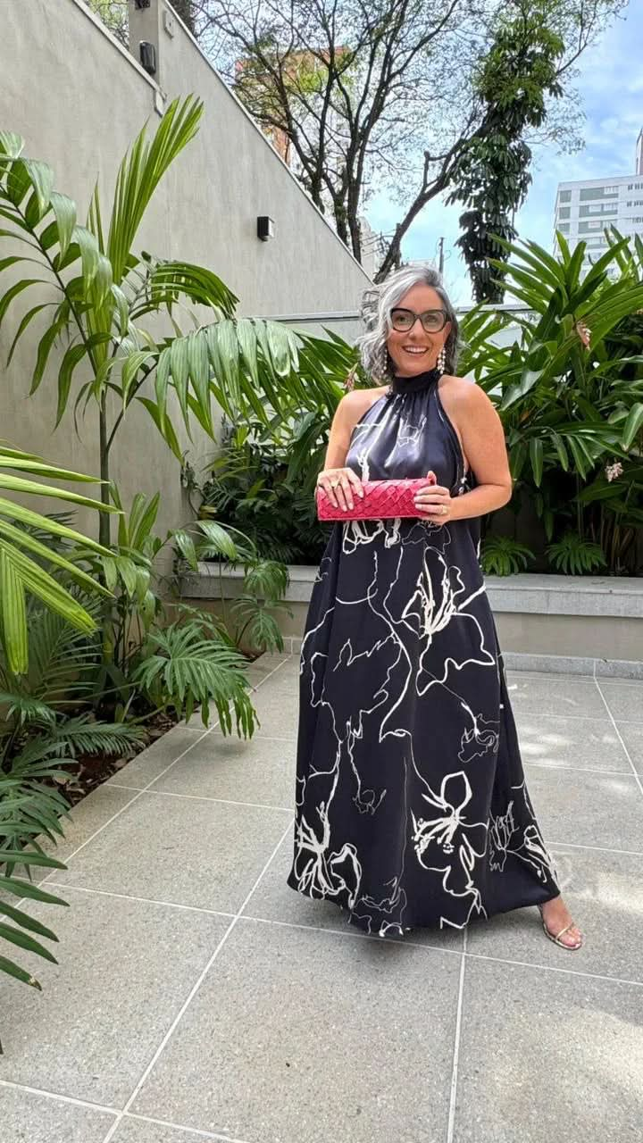 Mulher com vestido longo preto estampado, clutch vermelha, em jardim com plantas tropicais EZILDINHA