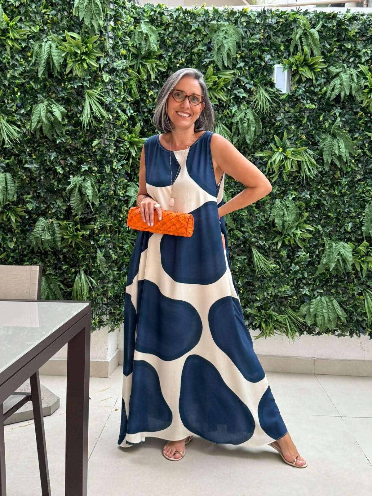 Mulher usando vestido regata longo com estampa de poá grande azul marinho, segurando clutch laranja, em área externa com parede de plantas EZILDINHA