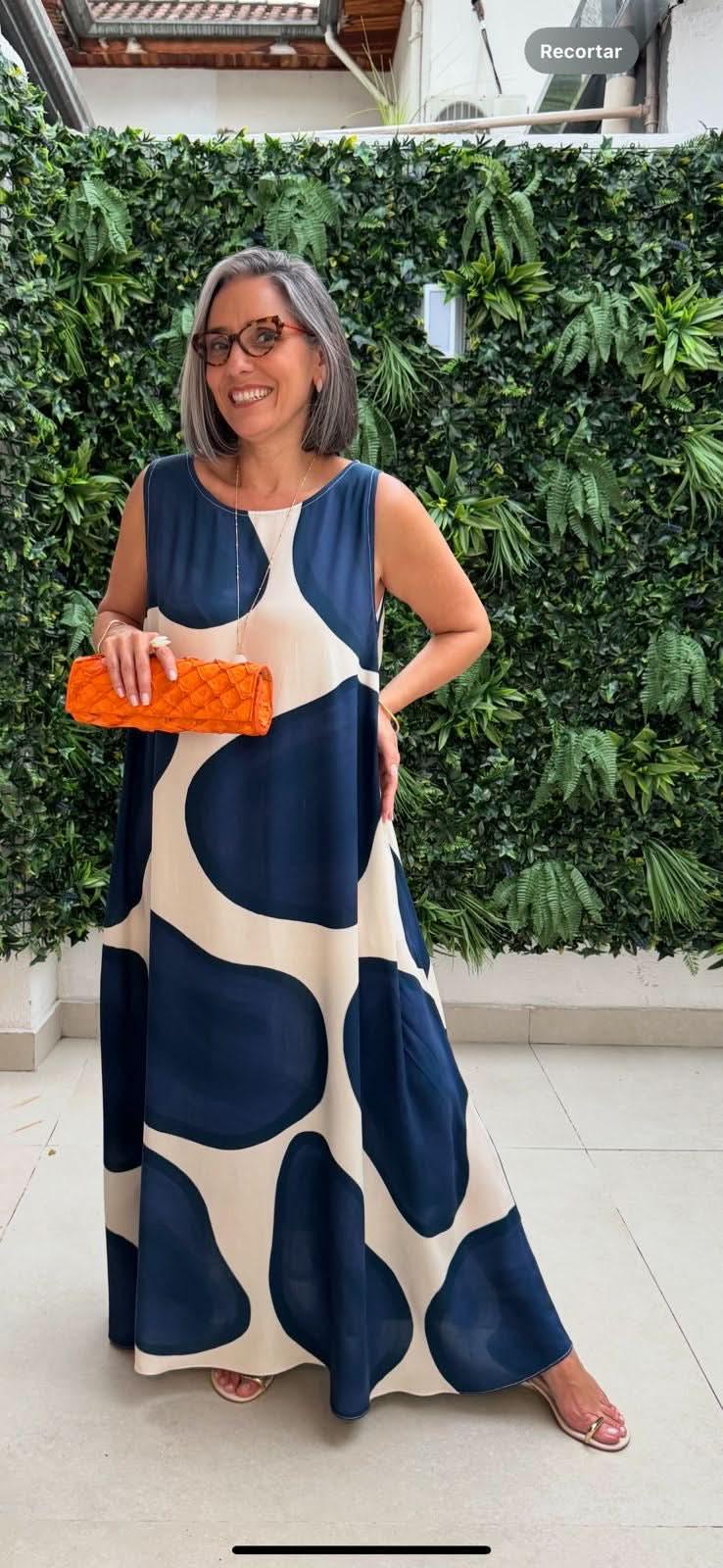 Mulher usando vestido longo regata com estampa de poá azul e bege, bolsa laranja e óculos, em ambiente com parede de plantas EZILDINHA