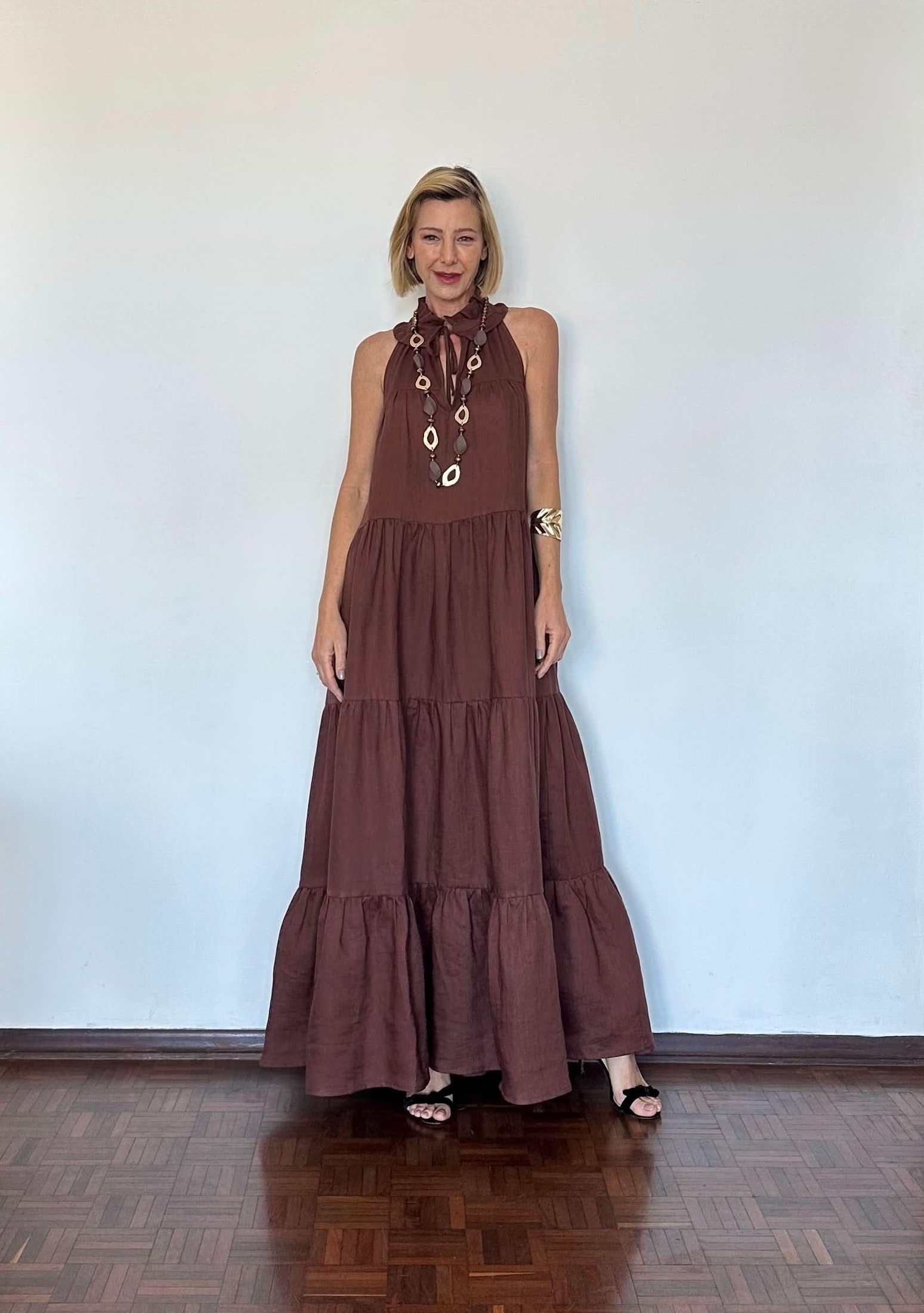 Mulher usando vestido longo de linho marrom, decote halter, com colar e sandália preta EZILDINHA