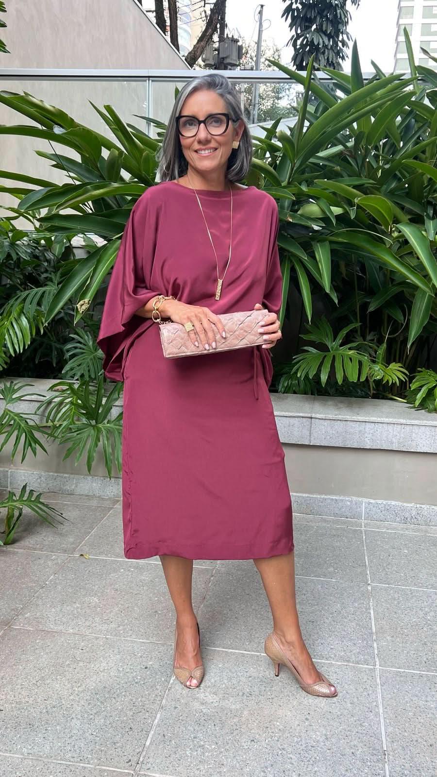 Mulher usando vestido midi de seda vinho, sapato nude e clutch bege, em ambiente externo com plantas EZILDINHA