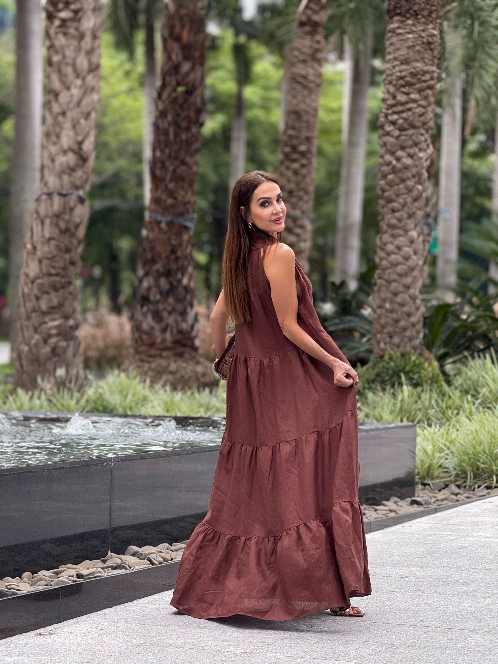 Mulher usando vestido longo de linho marrom ao ar livre, próximo a fonte e palmeiras EZILDINHA