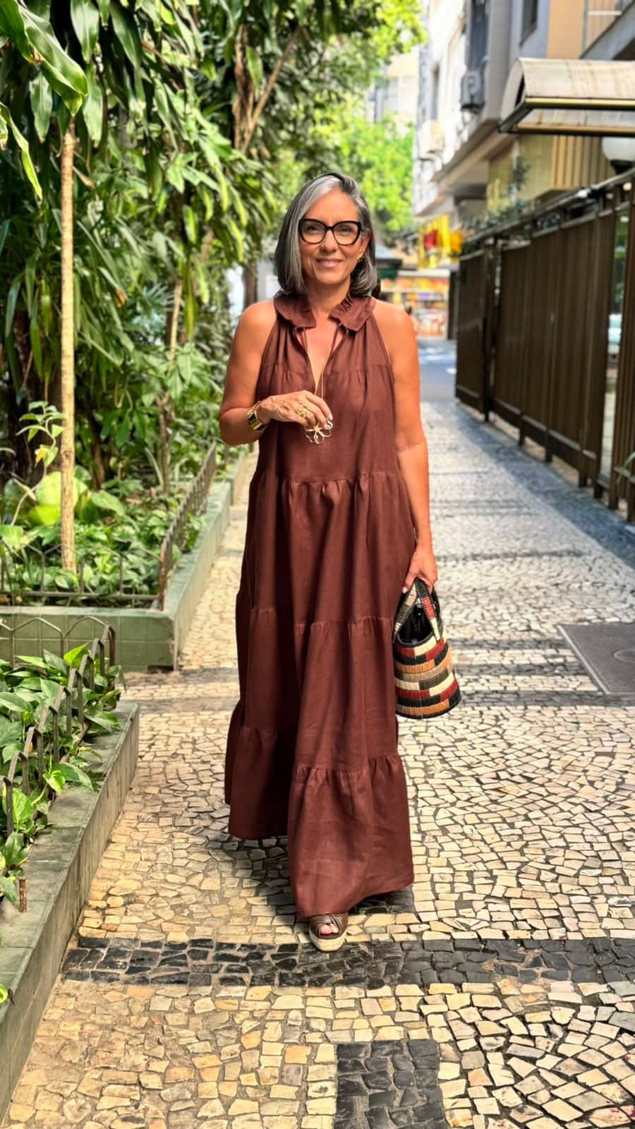 Mulher usando vestido longo de linho marrom, bolsa listrada, em calçada urbana arborizada EZILDINHA