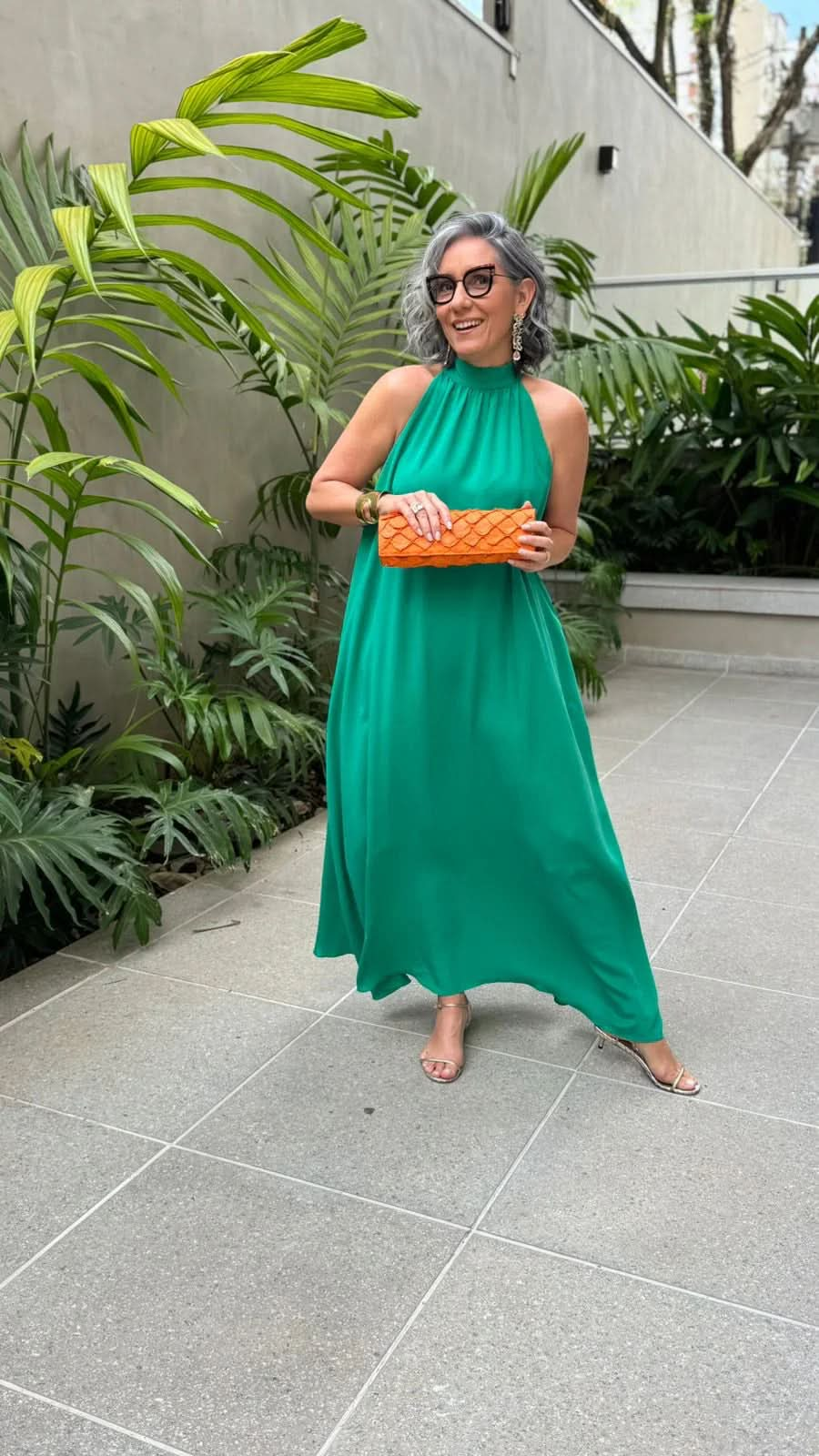 Mulher de cabelo grisalho usa vestido longo verde elegante, clutch laranja e sandália de salto, em área externa com plantas tropicais EZILDINHA