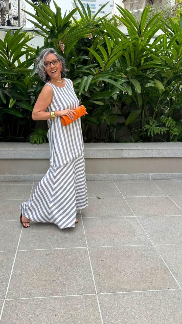 Mulher sorridente usando vestido longo listrado de linho, segurando clutch laranja, em jardim urbano EZILDINHA