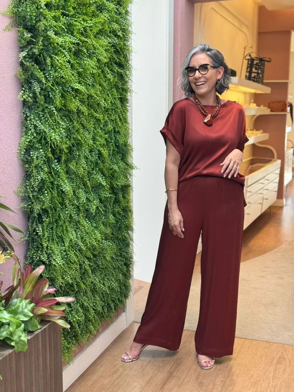 Mulher sorridente com cabelo grisalho curto usando conjunto de seda vinho, sandálias e colar marrom, em ambiente interno moderno com parede verde EZILDINHA