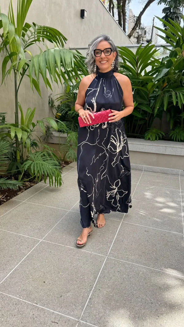 Mulher usando vestido longo estampado azul marinho de seda, segurando clutch rosa, em ambiente externo com plantas tropicais EZILDINHA
