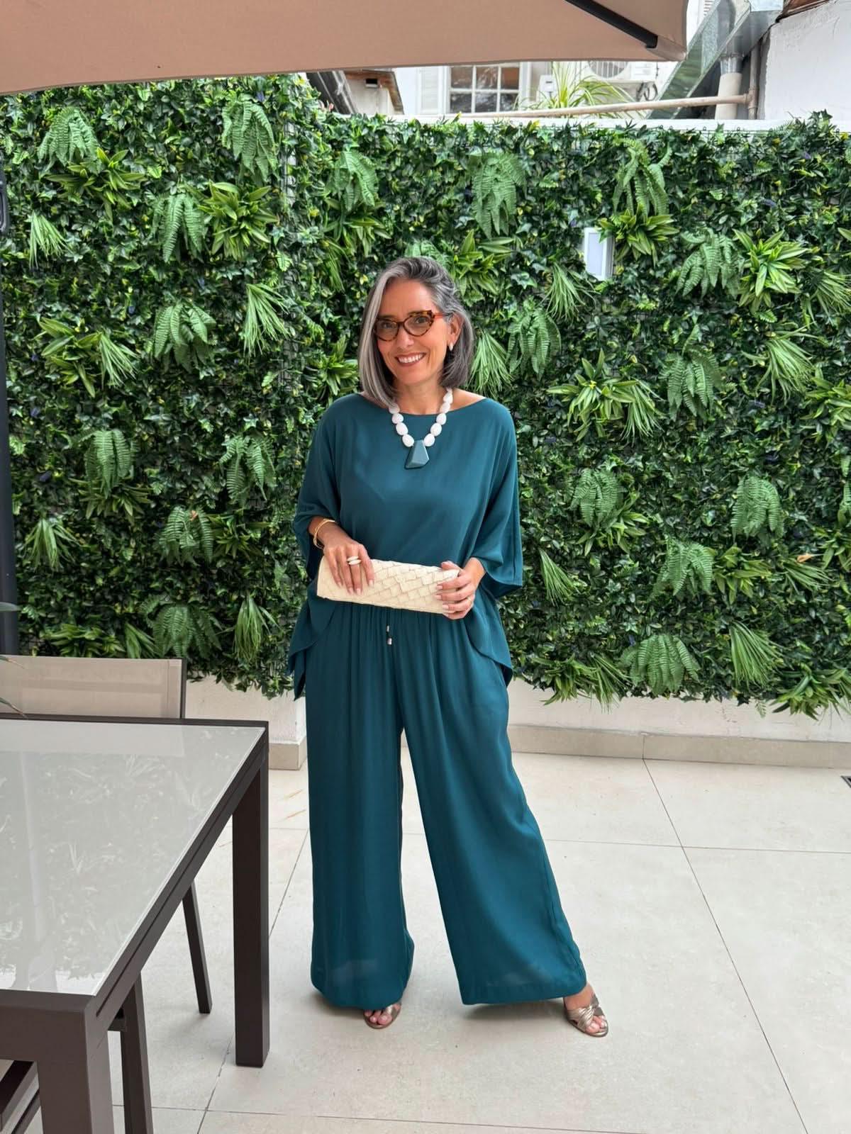 Mulher usando conjunto de blusa e calça azul em tecido leve, com bolsa clara, em ambiente externo com parede verde EZILDINHA