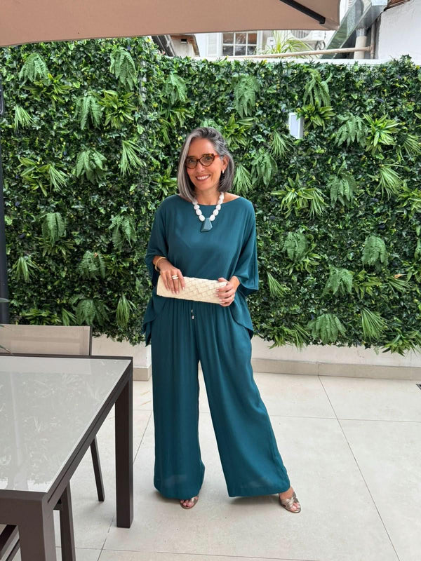 Mulher usando conjunto de blusa e calça azul em tecido leve, com bolsa clara, em ambiente externo com parede verde EZILDINHA