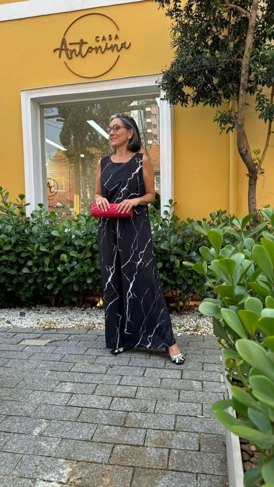 Mulher usando conjunto preto com estampa marmorizada branca, segurando clutch vermelha, em frente à fachada amarela com plantas EZILDINHA