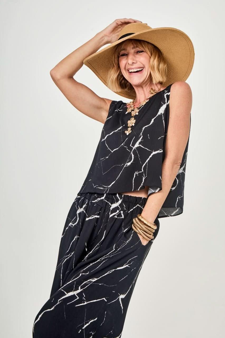 Mulher sorridente usando conjunto preto com estampa marmorizada, chapéu de palha e acessórios dourados EZILDINHA