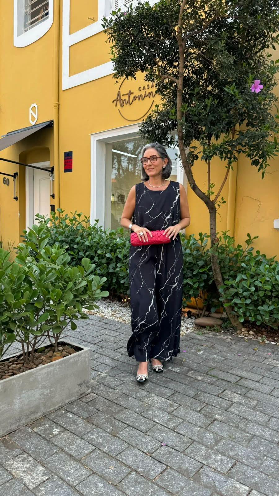 Mulher usando vestido longo preto com estampa marmorizada, segurando clutch rosa, em ambiente externo EZILDINHA