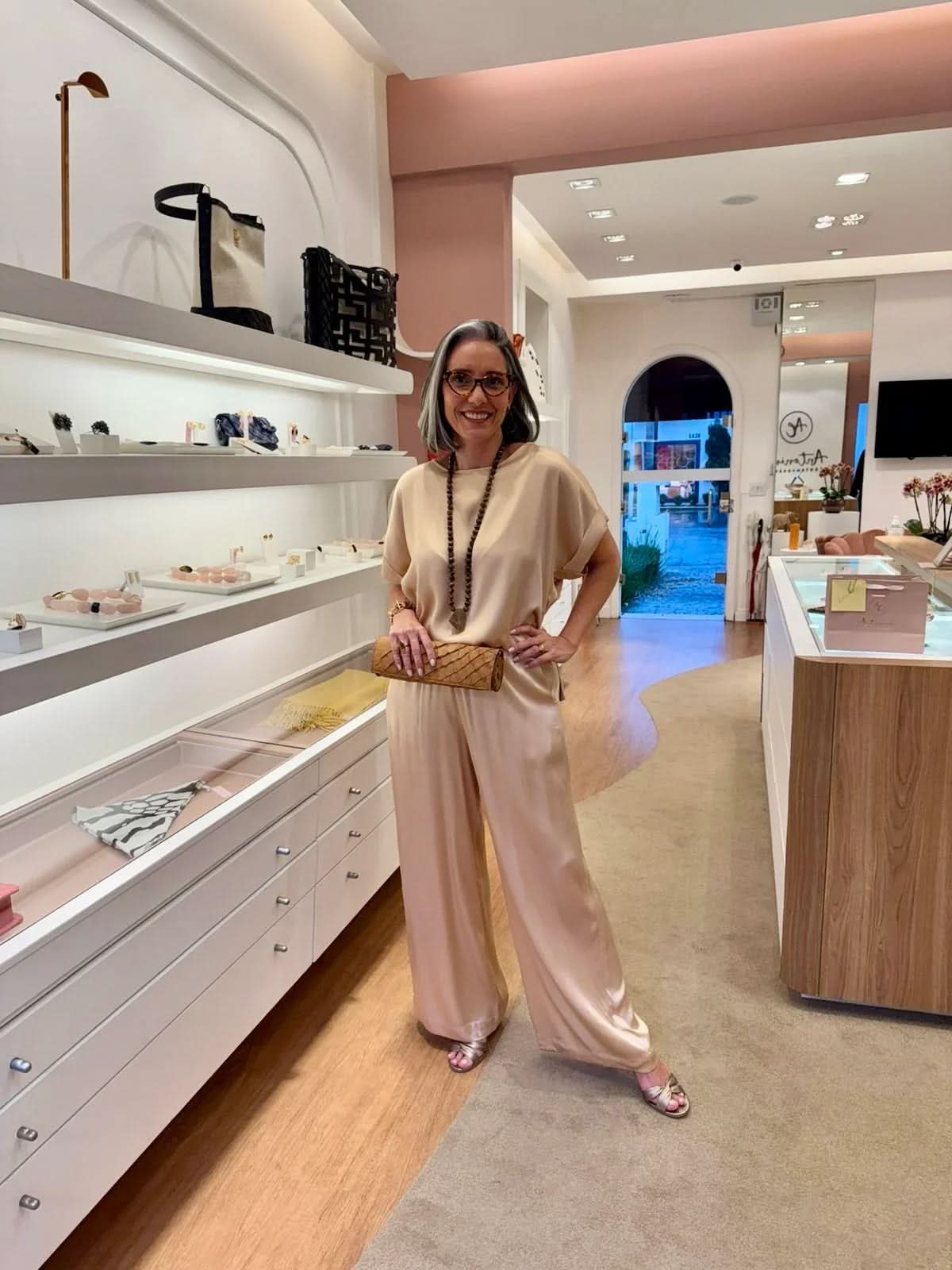 Mulher elegante com conjunto bege de seda, clutch e colar, em loja de moda feminina sofisticada EZILDINHA