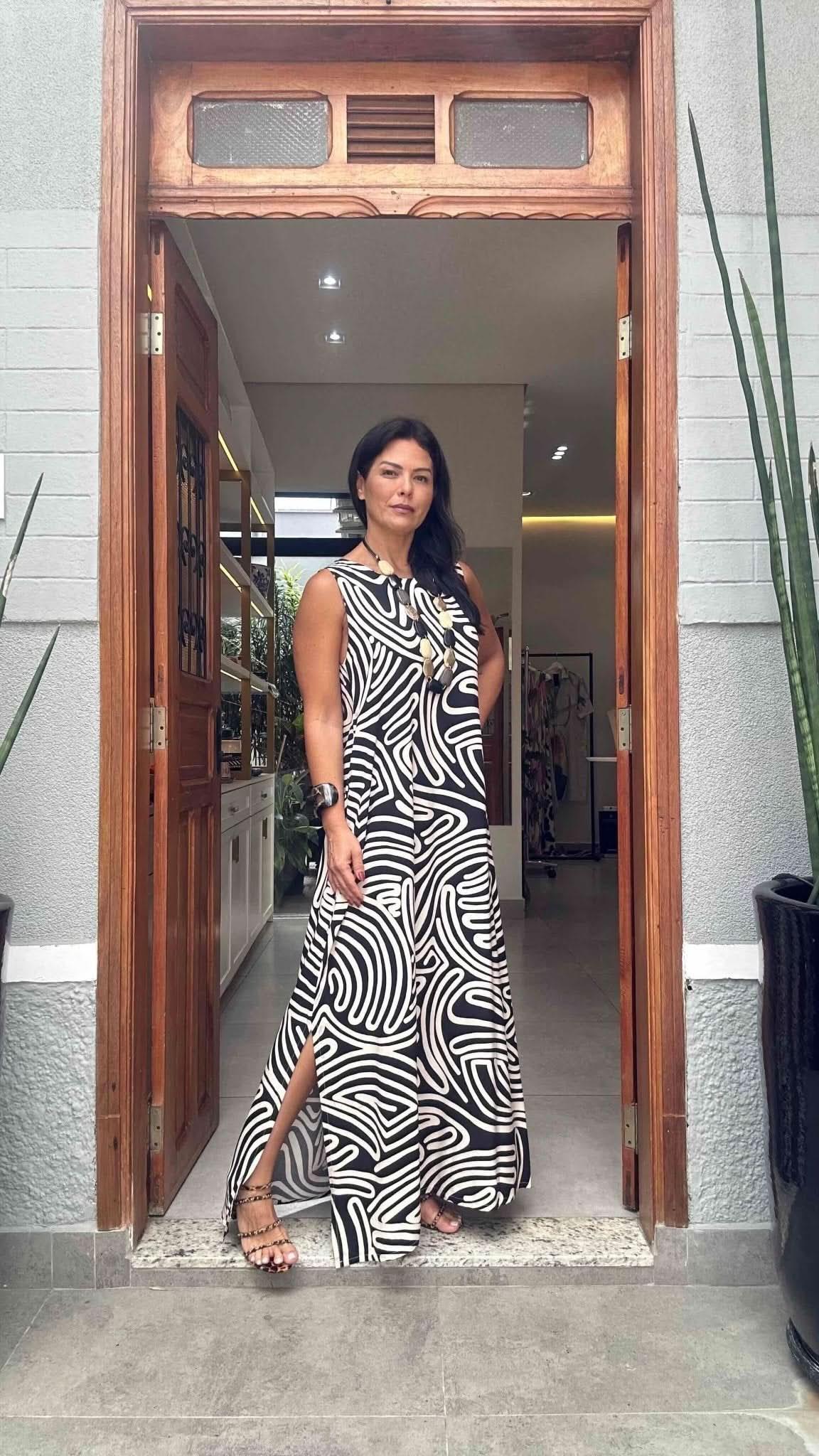 Mulher usando vestido longo preto e branco com estampa de linhas, em pé na porta de entrada EZILDINHA