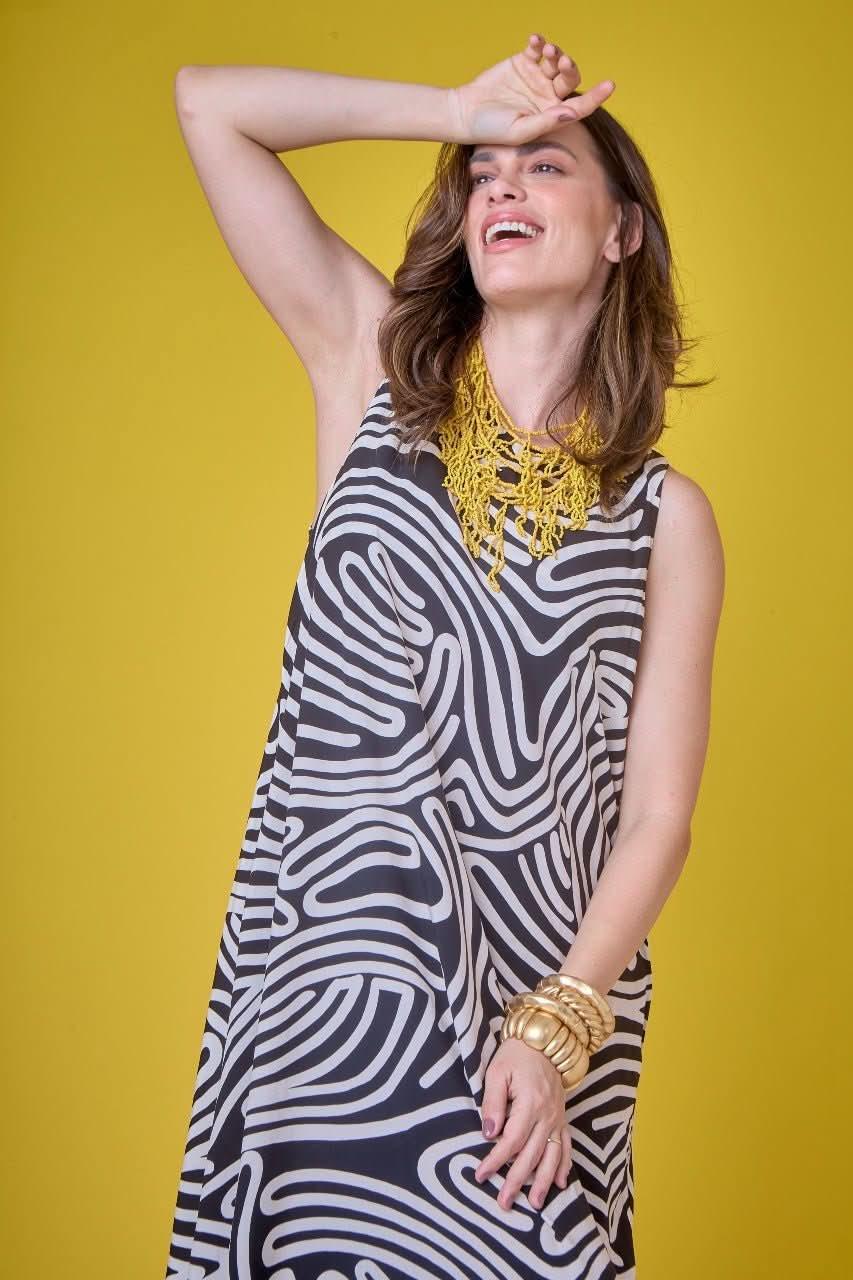 Mulher usando vestido midi preto e branco com estampa de linhas, acessórios dourados e colar amarelo, em fundo amarelo EZILDINHA