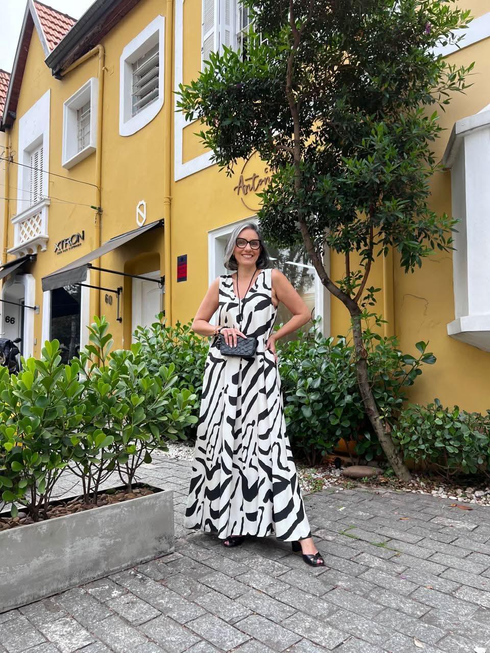 Mulher usando vestido longo branco com estampas pretas, em frente a casa amarela com plantas EZILDINHA