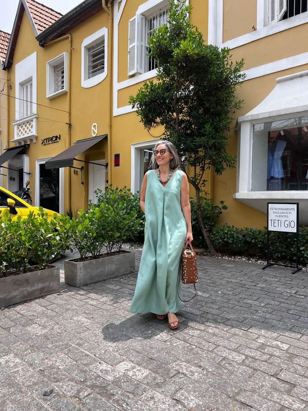 Mulher usando vestido longo verde de linho com pregas, andando na rua, em frente a casas amarelas EZILDINHA
