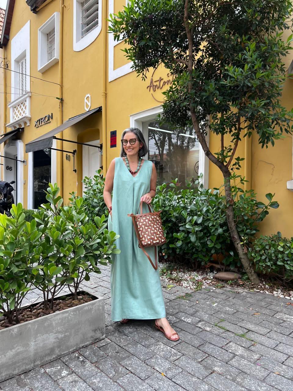 Mulher usando vestido longo de linho verde com pregas, bolsa estampada e sandálias, em rua arborizada EZILDINHA