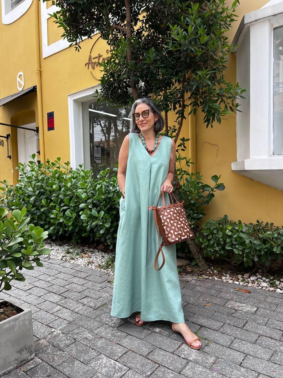 Mulher usando vestido longo de linho verde claro, bolsa de couro e colar, em área urbana com plantas EZILDINHA