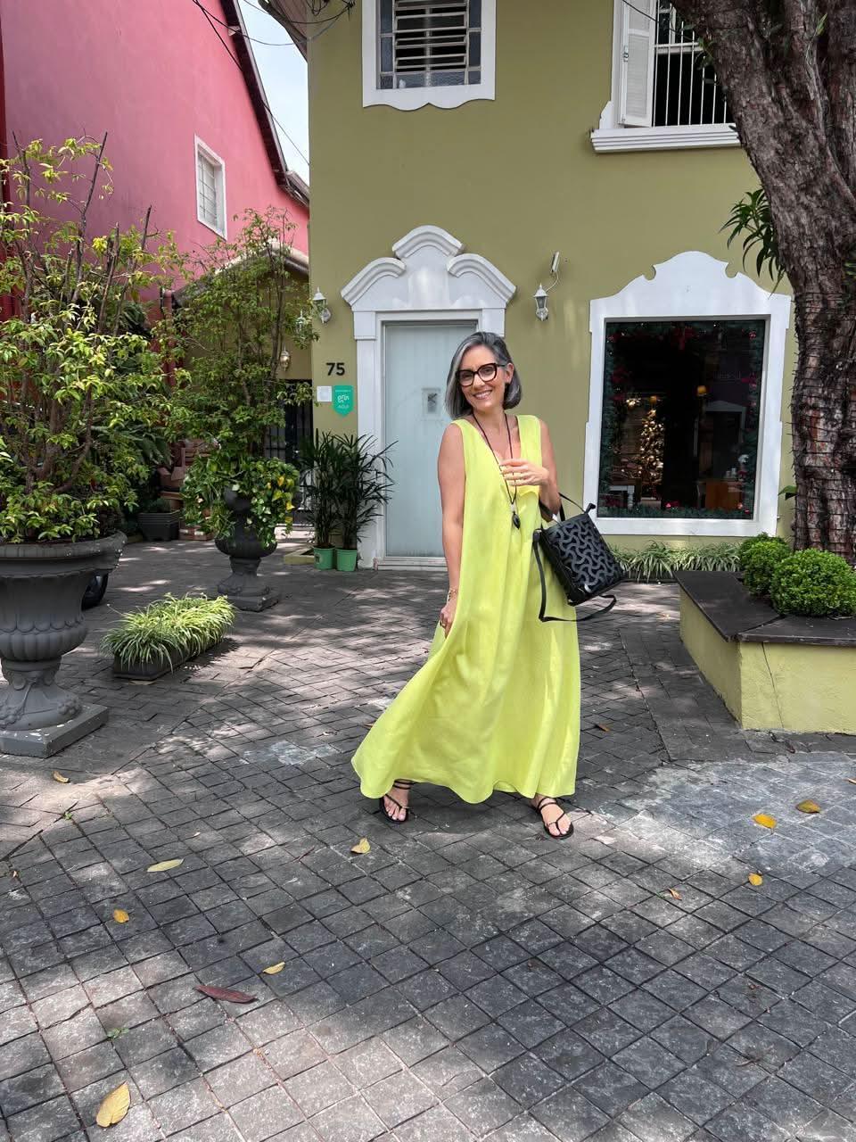 Mulher usando vestido de linho amarelo claro com pregas, óculos escuros e sandálias, em ambiente externo urbano arborizado EZILDINHA