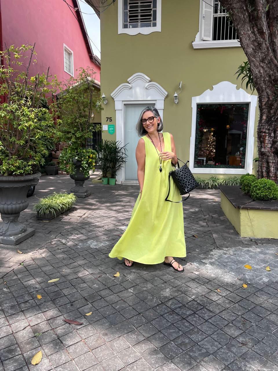 Mulher usando vestido longo de linho amarelo claro com pregas, ao ar livre em frente a uma casa decorada, segurando uma bolsa preta EZILDINHA