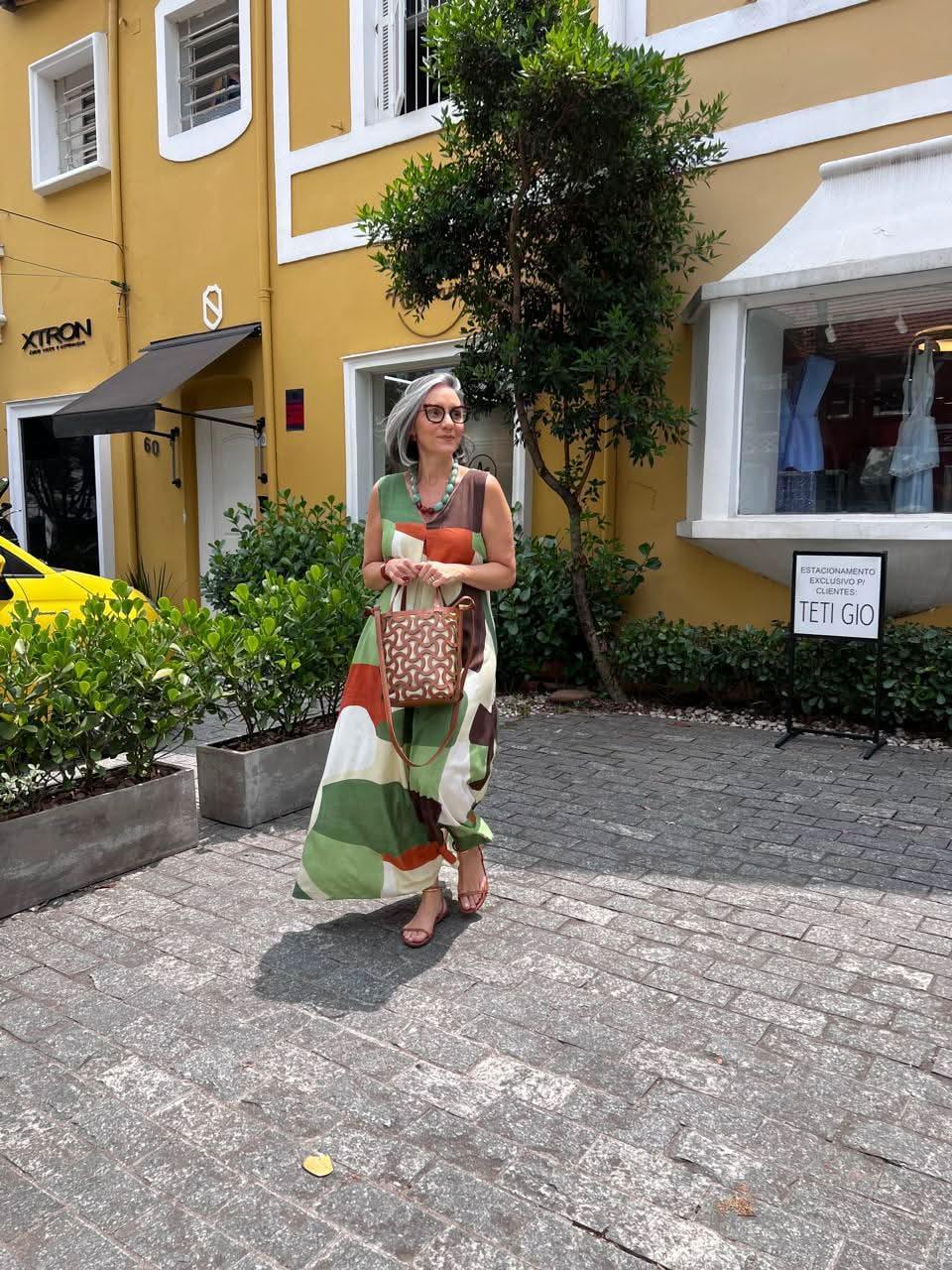 Mulher usando vestido longo colorido com estampas geométricas verdes, laranja e bege, sandálias e bolsa com padrão geométrico, caminhando em calçada urbana durante o dia EZILDINHA
