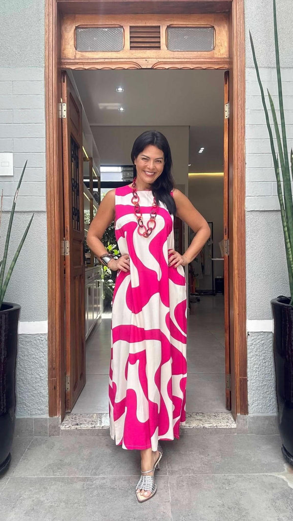 Mulher usando vestido longo estampado rosa e branco, em ambiente externo com plantas EZILDINHA
